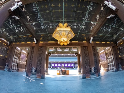 A traditional architectural interior features ornate wooden columns and beams adorned with intricate carvings. A large, decorative lantern with gold accents hangs from the ceiling. The floor is paved with polished stone tiles. In the background, there is a distant structure with purple and white decorations.