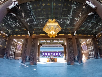 A traditional architectural interior features ornate wooden columns and beams adorned with intricate carvings. A large, decorative lantern with gold accents hangs from the ceiling. The floor is paved with polished stone tiles. In the background, there is a distant structure with purple and white decorations.