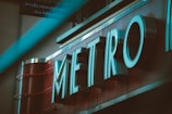 Large metallic letters spelling 'METRO' are prominently displayed with a teal lighting effect. The sign appears to be mounted on a red background, suggesting the entrance to a subway or a transportation hub. There's a blurred element in the foreground, adding depth to the image.