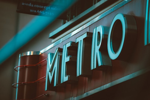 Large metallic letters spelling 'METRO' are prominently displayed with a teal lighting effect. The sign appears to be mounted on a red background, suggesting the entrance to a subway or a transportation hub. There's a blurred element in the foreground, adding depth to the image.