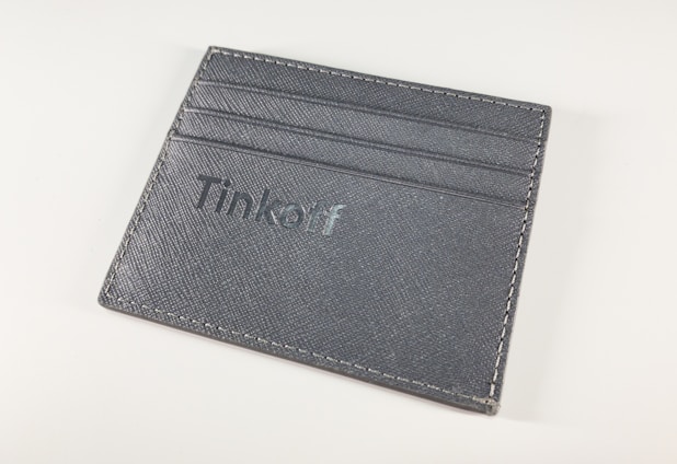 A gray leather cardholder features multiple slots for cards and has the word 'Tinkoff' embossed on the front. The stitching is visible around the edges, set against a plain white background.