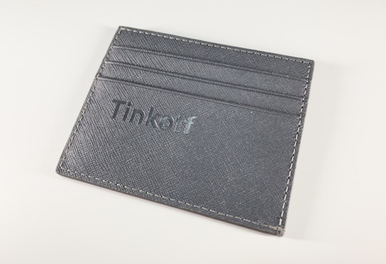 A gray leather cardholder features multiple slots for cards and has the word 'Tinkoff' embossed on the front. The stitching is visible around the edges, set against a plain white background.