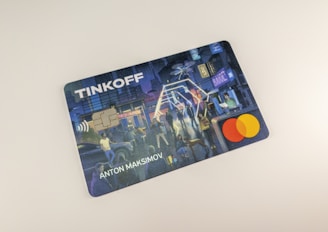 A credit card featuring a digital illustration on its surface. The scene depicts a bustling city environment at night with people engaging in various activities. Colorful lights and urban elements such as buildings and signs are visible. A contactless payment icon is printed above the embedded chip, and the Mastercard logo is displayed.