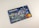 A credit card featuring a digital illustration on its surface. The scene depicts a bustling city environment at night with people engaging in various activities. Colorful lights and urban elements such as buildings and signs are visible. A contactless payment icon is printed above the embedded chip, and the Mastercard logo is displayed.