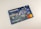 A credit card featuring a digital illustration on its surface. The scene depicts a bustling city environment at night with people engaging in various activities. Colorful lights and urban elements such as buildings and signs are visible. A contactless payment icon is printed above the embedded chip, and the Mastercard logo is displayed.