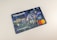 A credit card featuring a digital illustration on its surface. The scene depicts a bustling city environment at night with people engaging in various activities. Colorful lights and urban elements such as buildings and signs are visible. A contactless payment icon is printed above the embedded chip, and the Mastercard logo is displayed.