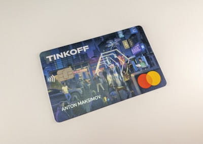 A credit card featuring a digital illustration on its surface. The scene depicts a bustling city environment at night with people engaging in various activities. Colorful lights and urban elements such as buildings and signs are visible. A contactless payment icon is printed above the embedded chip, and the Mastercard logo is displayed.