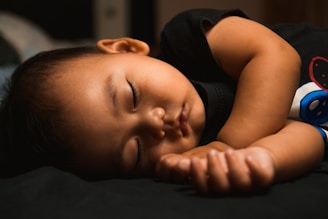 A young child is peacefully sleeping in a fetal position, wearing a black shirt with a colorful design. The lighting is soft and warm, highlighting the gentle contours of the child's face and hair.