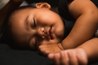 A peaceful baby is sleeping with eyes closed and a calm expression. The child is lying on a dark surface, with one arm resting near the face. The lighting is soft, highlighting the gentle features and serene atmosphere.