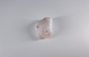 A smooth, oblong white stone with subtle brownish spots and markings lies centered on a plain, light gray surface.