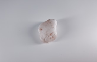 A smooth, oblong white stone with subtle brownish spots and markings lies centered on a plain, light gray surface.