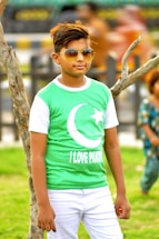 A young person wearing sunglasses and a green shirt featuring a crescent and star symbol paired with the words 'I Love Pakistan.' They are standing outdoors next to a tree, with a blurred background hinting at other people and greenery.