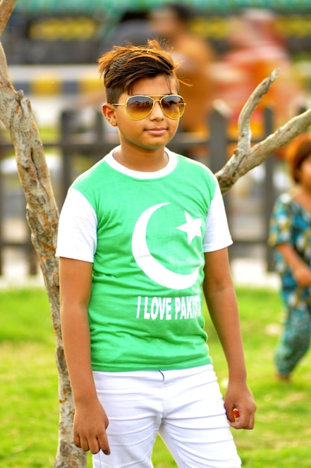 A young person wearing sunglasses and a green shirt featuring a crescent and star symbol paired with the words 'I Love Pakistan.' They are standing outdoors next to a tree, with a blurred background hinting at other people and greenery.