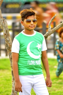 A young person wearing sunglasses and a green shirt featuring a crescent and star symbol paired with the words 'I Love Pakistan.' They are standing outdoors next to a tree, with a blurred background hinting at other people and greenery.