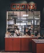A kitchen scene with chefs working diligently behind a counter. The area is equipped with professional kitchen appliances and is marked by a sign that reads 'CUCINA'. The counter features various items such as dishes, a potted plant, and kitchen utensils. Warm lighting adds a cozy atmosphere to the environment.