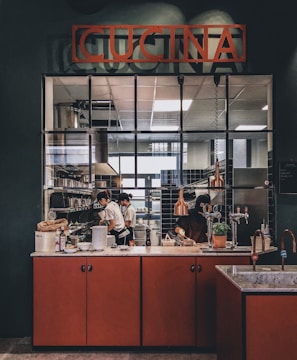A kitchen scene with chefs working diligently behind a counter. The area is equipped with professional kitchen appliances and is marked by a sign that reads 'CUCINA'. The counter features various items such as dishes, a potted plant, and kitchen utensils. Warm lighting adds a cozy atmosphere to the environment.