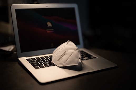A laptop is placed on a dark surface with a face mask resting on its keyboard. The laptop screen displays a dimmed login screen, adding to the subdued lighting of the scene.