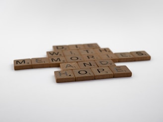 Wooden Scrabble tiles arranged on a white surface forming the words 'HOPE', 'MEMORIES', 'AND', 'WITH', and 'DIE' in a crossword style.