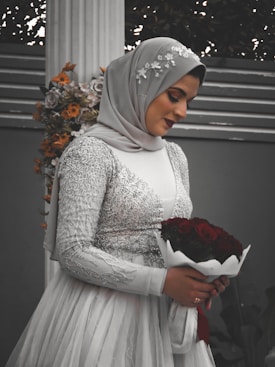 A woman wearing an embellished silver hijab and embroidered gown, holding a bouquet of red roses. The background includes a floral arrangement with orange and white flowers against a grey wall.