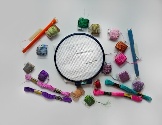 An arrangement of colorful embroidery thread skeins encircles a circular embroidery hoop with a white cloth. The threads are in various shades including pink, purple, green, orange, and blue. Several bobbins of thread are visible, as well as two embroidery needles placed on the cloth within the hoop.