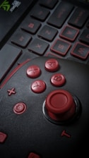 A close-up of a gaming joystick with red buttons marked A, B, X, and Y. The joystick is placed on top of a laptop keyboard, with keys outlined in red. The image is dimly lit, enhancing the contrast between the red components and the dark background.