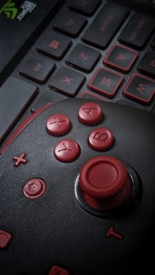 A close-up of a gaming joystick with red buttons marked A, B, X, and Y. The joystick is placed on top of a laptop keyboard, with keys outlined in red. The image is dimly lit, enhancing the contrast between the red components and the dark background.