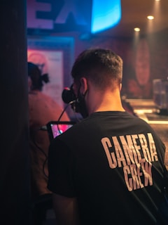 A person wearing a 'CAMERA CREW' shirt is operating a camera in a dimly lit indoor setting, possibly a studio or a venue. The background features blurred elements, with a soft blue and pink color palette.