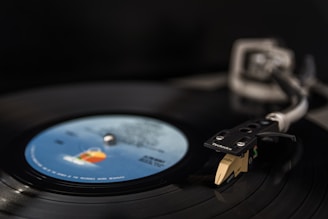 A close-up view of a vinyl record playing on a turntable. The tonearm and needle are positioned on the record, which features a colorful label in the center. The lighting creates a soft, focused effect on the needle and label, with a dark, blurred background.