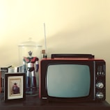A vintage television set with knobs and an antenna is placed on a dark wooden surface. Beside it, a framed photograph of a child dressed in a cowboy outfit stands in front of a row of books. A large robot figurine with red and black colors is encased in a glass jar, positioned to the left of the TV set.