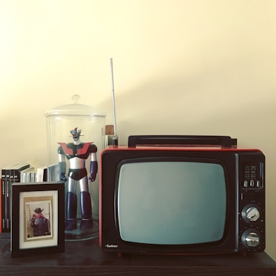 A vintage television set with knobs and an antenna is placed on a dark wooden surface. Beside it, a framed photograph of a child dressed in a cowboy outfit stands in front of a row of books. A large robot figurine with red and black colors is encased in a glass jar, positioned to the left of the TV set.