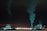 An industrial complex is prominently featured at night, illuminated with bluish-green lights. Large chimneys expel smoke into the dark sky, and several wind turbines are visible in the background, suggesting a contrast between industrial production and renewable energy sources.