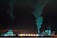 An industrial complex is prominently featured at night, illuminated with bluish-green lights. Large chimneys expel smoke into the dark sky, and several wind turbines are visible in the background, suggesting a contrast between industrial production and renewable energy sources.