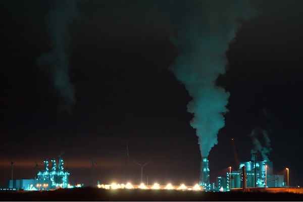 An industrial complex is prominently featured at night, illuminated with bluish-green lights. Large chimneys expel smoke into the dark sky, and several wind turbines are visible in the background, suggesting a contrast between industrial production and renewable energy sources.