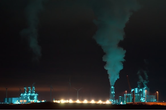 An industrial complex is prominently featured at night, illuminated with bluish-green lights. Large chimneys expel smoke into the dark sky, and several wind turbines are visible in the background, suggesting a contrast between industrial production and renewable energy sources.