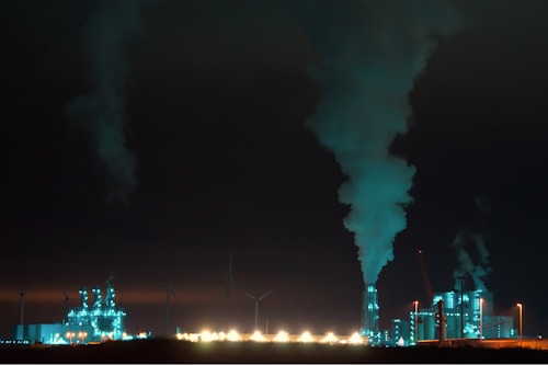 An industrial complex is prominently featured at night, illuminated with bluish-green lights. Large chimneys expel smoke into the dark sky, and several wind turbines are visible in the background, suggesting a contrast between industrial production and renewable energy sources.
