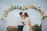 A bride and groom sharing a joyful kiss under a blooming archway at sunset.