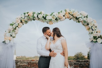 A bride and groom sharing a joyful kiss under a floral arch at sunset.