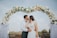 Bride and groom smiling under elegant pastel floral arch at outdoor wedding.