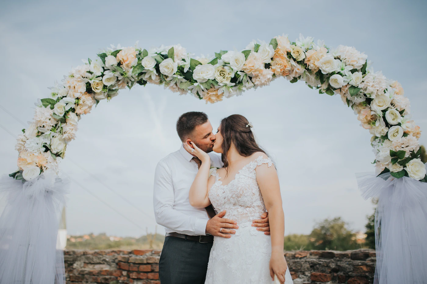 A bride and groom sharing their first kiss as a married couple under a round floral arch decorated with white and peach roses, with a stone brick wall visible behind them