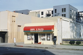 A small Mexican restaurant with a red and green awning, displaying the name 'Chips 'N Salsa'. The building is situated on a street corner, featuring a yellow exterior with windows covered in metal bars. Surrounding buildings are visible in the background, with a mix of architectural styles.