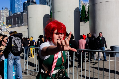 A person in vibrant cosplay attire stands in front of gray structures and a crowd of people. The individual has bright red hair and is wearing a green and white outfit with a checked scarf. Their hand is extended in a dynamic gesture. The background includes a cityscape with tall buildings and a clear blue sky.