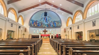 A spacious church interior with wooden pews arranged in rows leading up to an altar. Above the altar is a large, colorful mural depicting religious figures. The church has high arched ceilings with wooden beams and natural light streaming in through the arched windows on the sides.