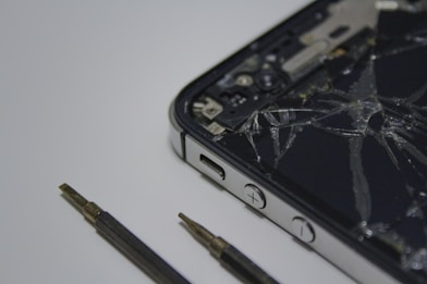 A broken smartphone with a cracked screen is placed on a surface, alongside a couple of small screwdrivers. The phone is partially disassembled and the internal components are visible.