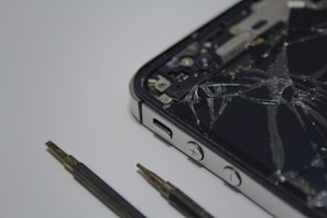 A broken smartphone with a cracked screen is placed on a surface, alongside a couple of small screwdrivers. The phone is partially disassembled and the internal components are visible.