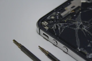 A broken smartphone with a cracked screen is placed on a surface, alongside a couple of small screwdrivers. The phone is partially disassembled and the internal components are visible.