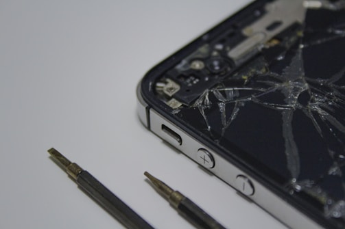 A broken smartphone with a cracked screen is placed on a surface, alongside a couple of small screwdrivers. The phone is partially disassembled and the internal components are visible.