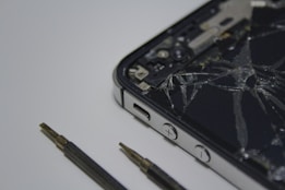 A broken smartphone with a cracked screen is placed on a surface, alongside a couple of small screwdrivers. The phone is partially disassembled and the internal components are visible.