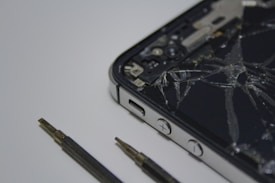 A broken smartphone with a cracked screen is placed on a surface, alongside a couple of small screwdrivers. The phone is partially disassembled and the internal components are visible.