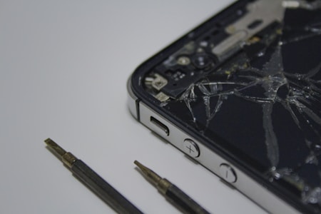 A broken smartphone with a cracked screen is placed on a surface, alongside a couple of small screwdrivers. The phone is partially disassembled and the internal components are visible.
