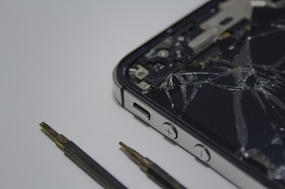 A broken smartphone with a cracked screen is placed on a surface, alongside a couple of small screwdrivers. The phone is partially disassembled and the internal components are visible.